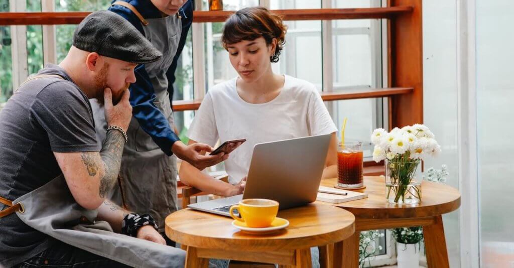 Frau an rundem Holztisch mit Laptop, ein Mann hinter ihr, der ein Handy zeigt und ein weitere mann neben ihr, der auf das Handy schaut